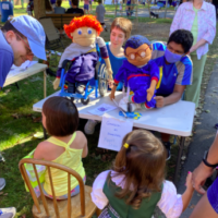 Two children putting on a puppet show for other children. One puppet is in a wheelchair and the other has a white cane such as used by some blind people.