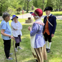 Two women in modern clothes and a man and woman in colonial clothing stand chatting in a cemetery, with U.S. flags in the background.