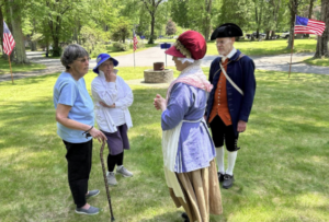 Two women in modern clothes and a man and woman in colonial clothing stand chatting in a cemetery, with U.S. flags in the background.