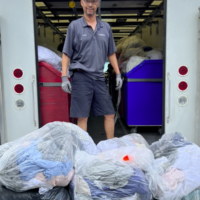 Photograph of man standing in the open back of a truck with a large pile of contributed linens in bags at his feet.