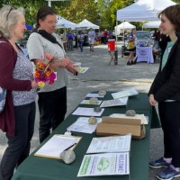 Attendees at a Farmers Market stand before informational paperwork on a table.