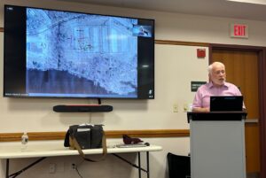 A speaker behind a podium and an aerial terrain map on a video screen.