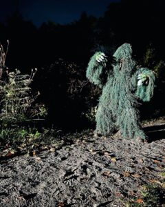 A 'monster' figure in a green ghillie suit against a dark background