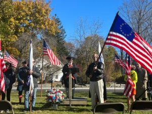 Aman holds an early American flag; a speakar at a podium and other flag-holders stand behind him