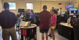 Adults and children mingle informaly with uniformed police officers in a room with multiple computer displays