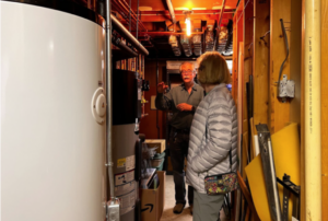 Man and woman in a basement with large tank and heating equipment. Man gesturing.