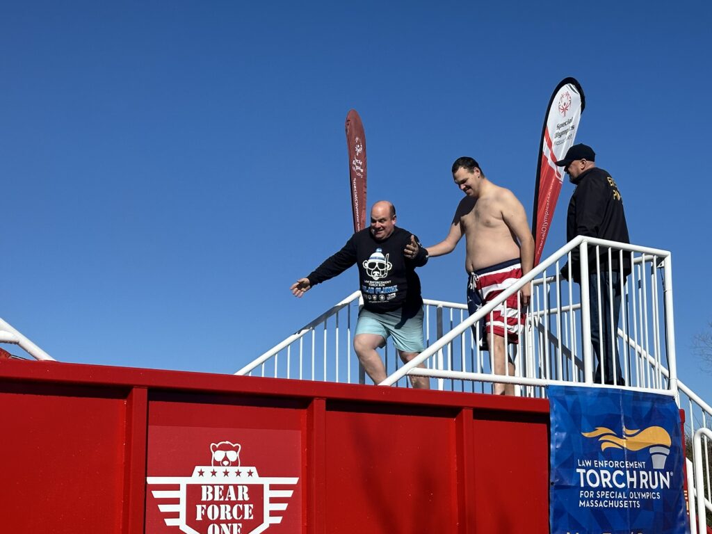 Large, red, rectangular metal tank with white letters saying, “Bear Force One” on the side and white railings at the top with two men in bathing suits moving toward the tank.