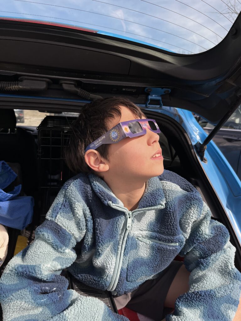 Boy wearing eclipse glasses looking up at the sky out of a car window.