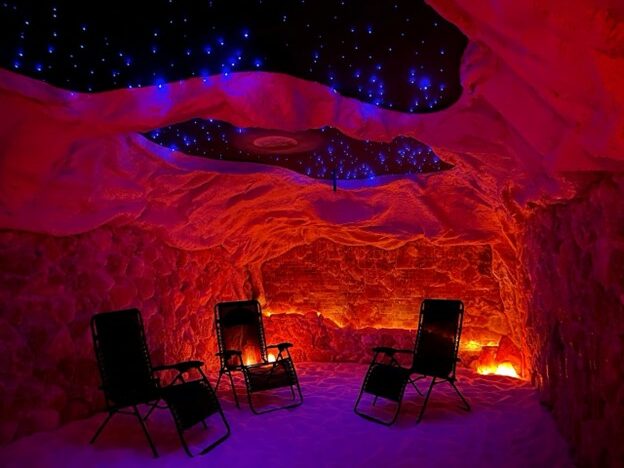 A dark, but colorful room with craggy, salt-covered, walls, stars on the ceiling, and orange lighting. There are several reclining chairs for a comfortable experience.