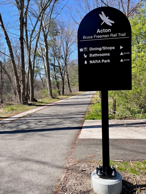 A black metal sign on the Bruce Freeman Rail Trail that shows distances to dining and shops, bathrooms, and NARA park.