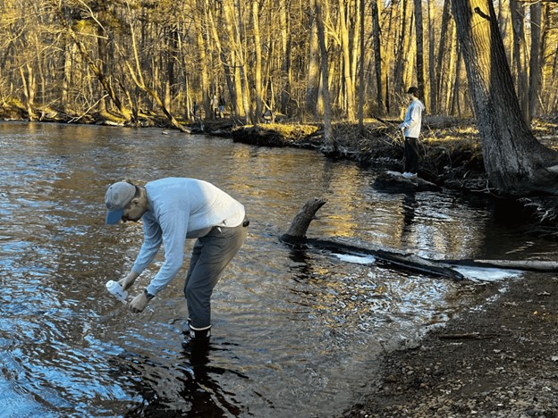 Man standing in ankle-deep water showing how to fill a water bottle for sampling. A person inthe background is fishing from a rock in the water, and the sun shines on trees along the shore.