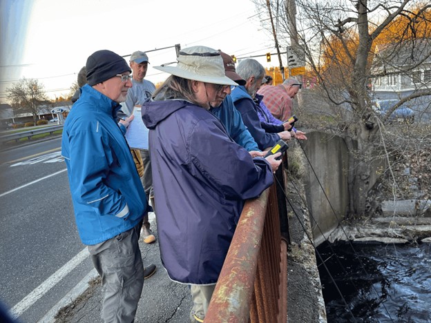 A group of people standing on an old bridge. Some of them are lowering instruments into the water.