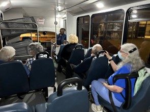 A woman stands at the front of bus full of riders and talks to the passengers.