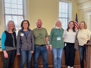 A group of six people standing in front of the windows at Town Hall Room 204.