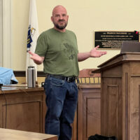 A bald, bearded, man stands in front of a podium. He is wearing jeans and a t-shirt, as befits a gardening expert.