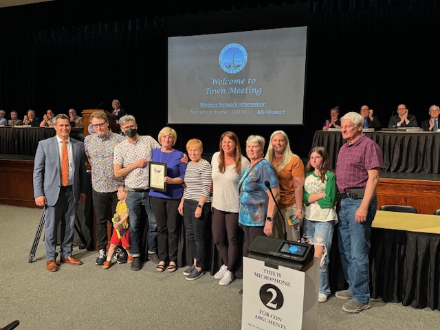 A group of people of various ages, including the Town Manager, Lalli Award recipients, and their families stand in front of Town Meeting.