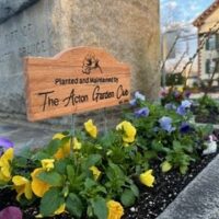 A cheery flowerbed with a wooden sign that says "Planted and maintained by the Acton Garden Club."