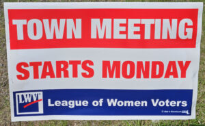 Red, white, and blue sign that says Town Meeting starts Monday. The sign is on a grassy area so it can be seen from the road.