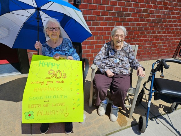 Two older women seated in the sun. One has an umbrella in one hand and a poster in the other that says "Happy 90's. Wishing you much happiness, good health, and lots of love always!"