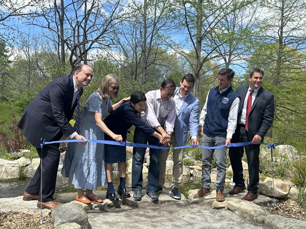 A line of people standing outside in front of a blue ribbon. Two people are cutting the ribbon.