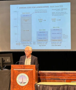 A grey-haired man in a suit jacket stands at a podium. A large screen is behind him with a page from his presentation on leaf blowers.