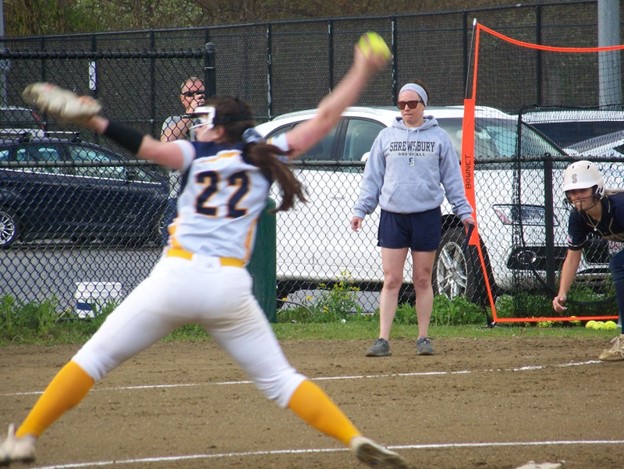 A woman in a white uniform has her arms spread wide as she winds up for a pitch.