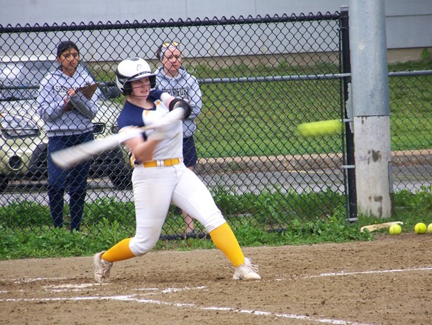 A young woman in a white uniform swings a bat as a ball comes in fast.