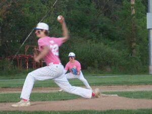 Teenaged boy in a baseball uniform throws a pitch. Another baseball player is ready to catch in the background.