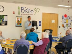 A man wearing a button-down shirt with rolled=up sleeves stands in front of the Acton Senior Center Bistro 30 sign. He is speaking to an audience of older citizens.