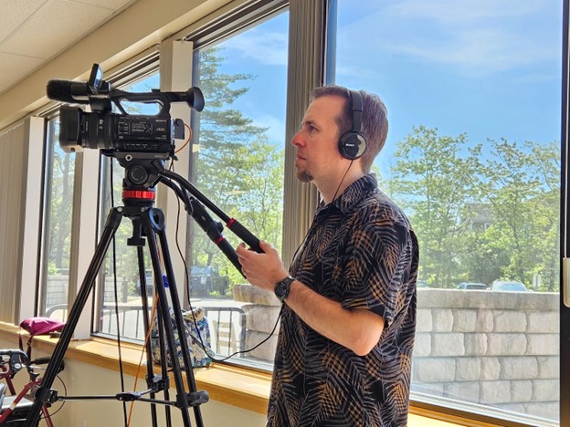 A man wearing a print shirt and headphones manages a large camera on a tripod while recording Java with John for ActonTV.