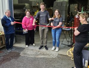 Five people stand in front of a shop located in an industrial complex. Two people hold a red ribbon and the new owners of Marzae Winery start to cut the ribbon with a very large pair of scissors.You can see large metal tanks and other wine-making parephernelia in the background.