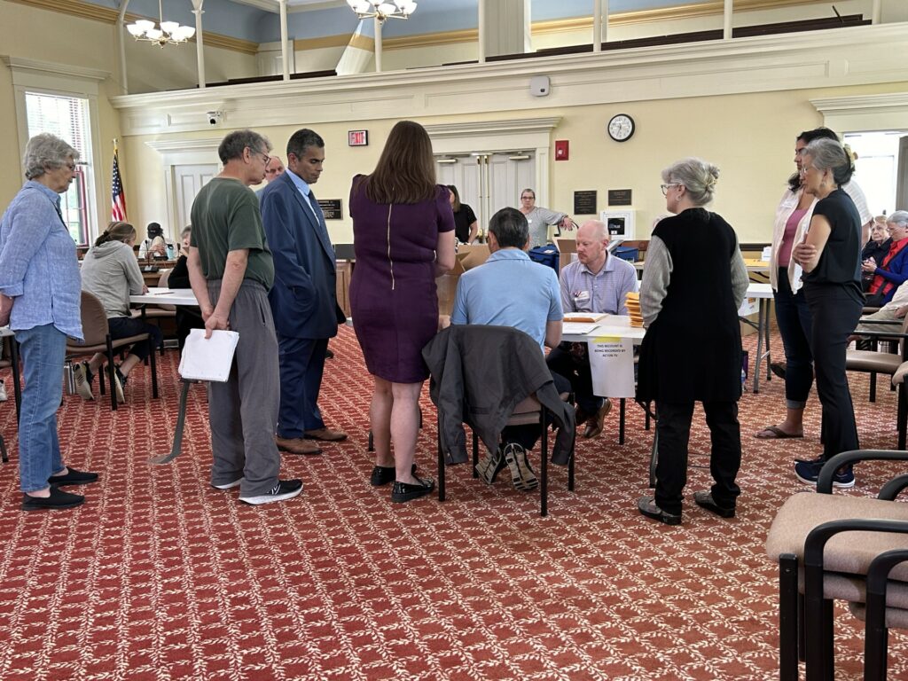 Three people are sitting at a table looking at something (probably a ballot). A number of people, including the Assistant Town Clerk, are huddled around the table, watching closely.