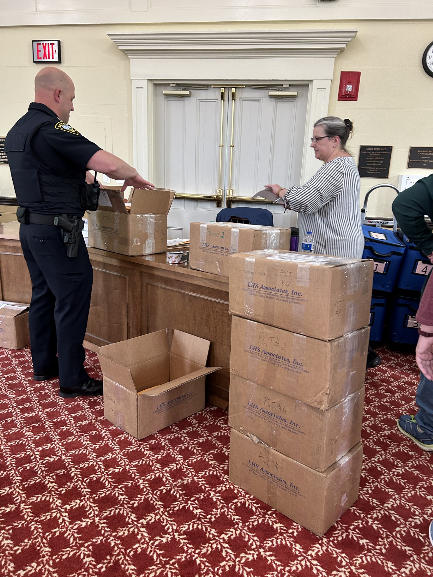 A police detective and a woman stand near a large stack of sealed boxes. The officer is opening one box that has been moved to a desk.