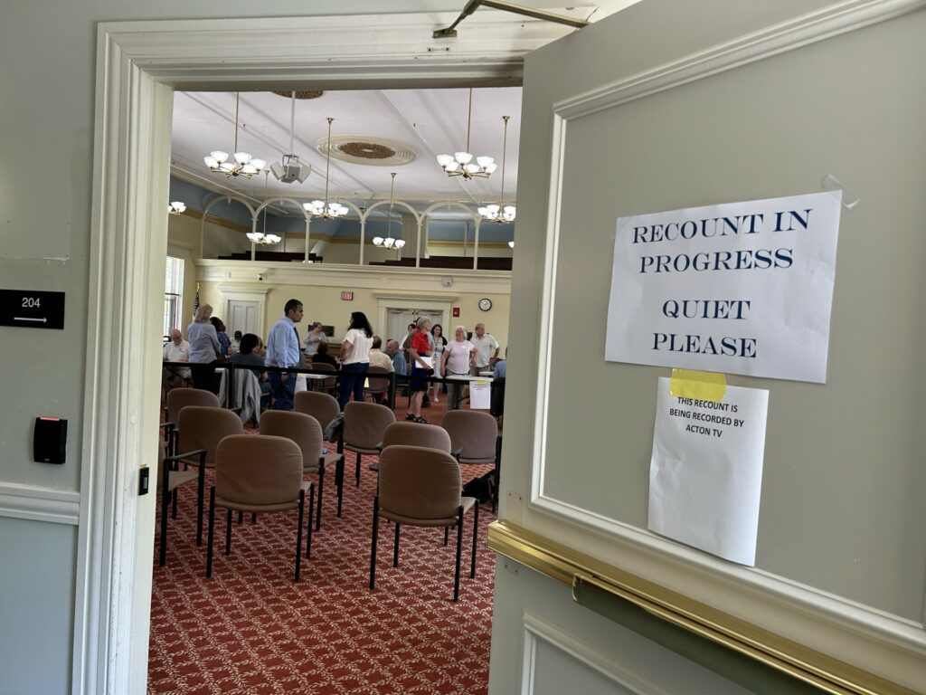 An open door with a view into a large room. Signs on the door say "Quiet Please, Recount in Progress," and "This recount is bing recorded by Acton TV." In the room, people are milling around. There are chairs set up for observers.