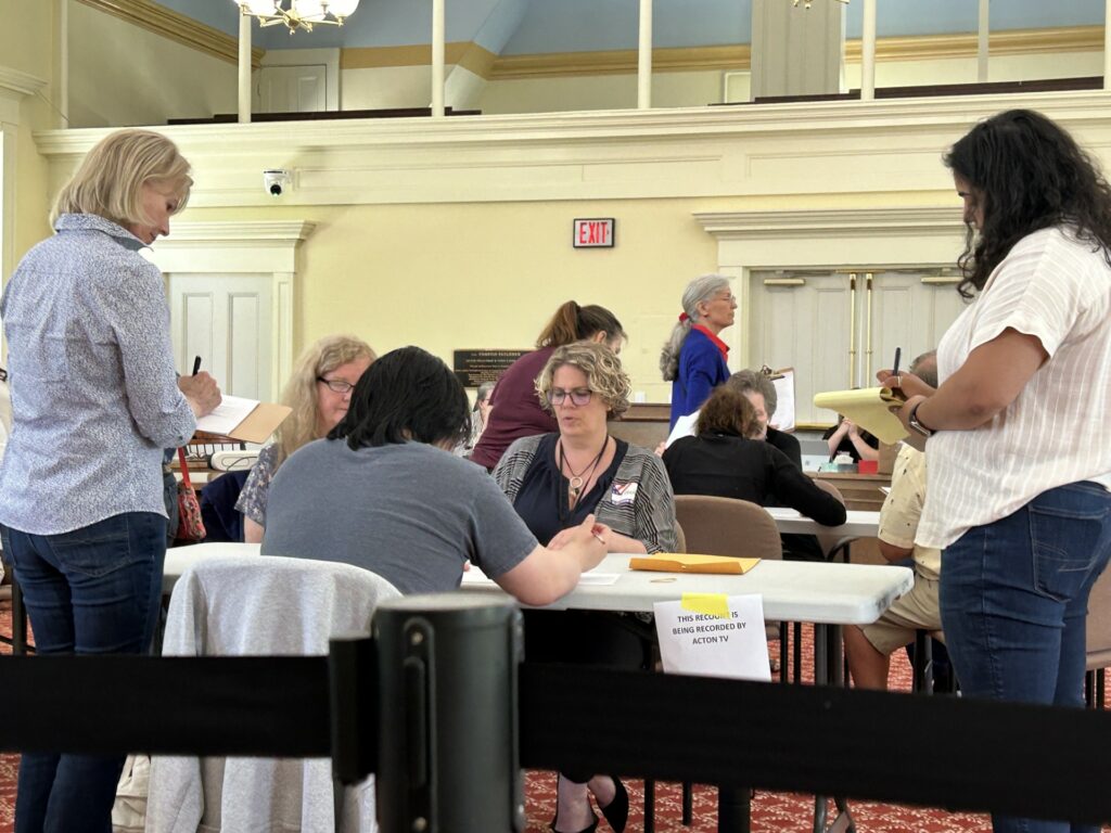 Three workers sit at a table counting the ballots. Two women stand nearby with clipboards, keeping a careful eye on the tallied ballots.