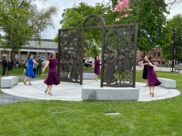 A group of women dancing on the plaza that surrounds the gate-like sculpture.