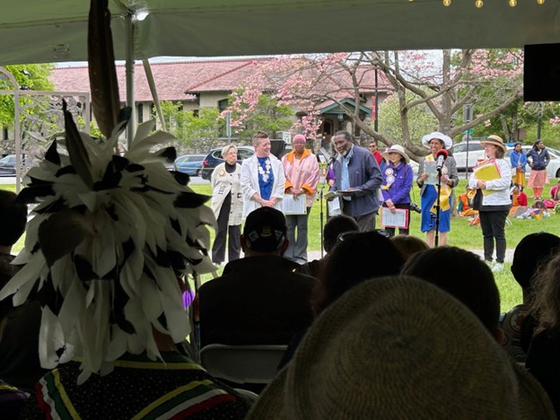 A Black man speaks to an audience on Lexington Green. A group of women stands in the background and someone wearing a feathered hat listens from the audience.