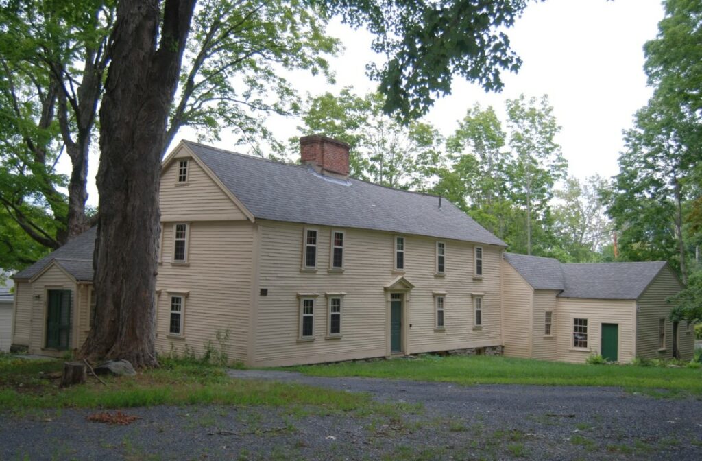 A Revolutionary-era white house with a center chimney with a large tree in the side yard. There are at least two additions to the original building.