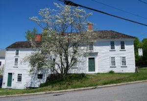 A Revolutionary-era white house with a center chimney. The main house is set on a hill and an ell on the left side has a small door that opens at a lower level.