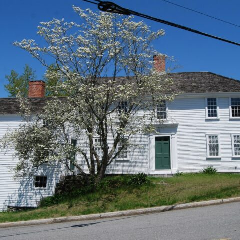 A Revolutionary-era white house with a center chimney. The main house is set on a hill and an ell on the left side has a small door that opens at a lower level.