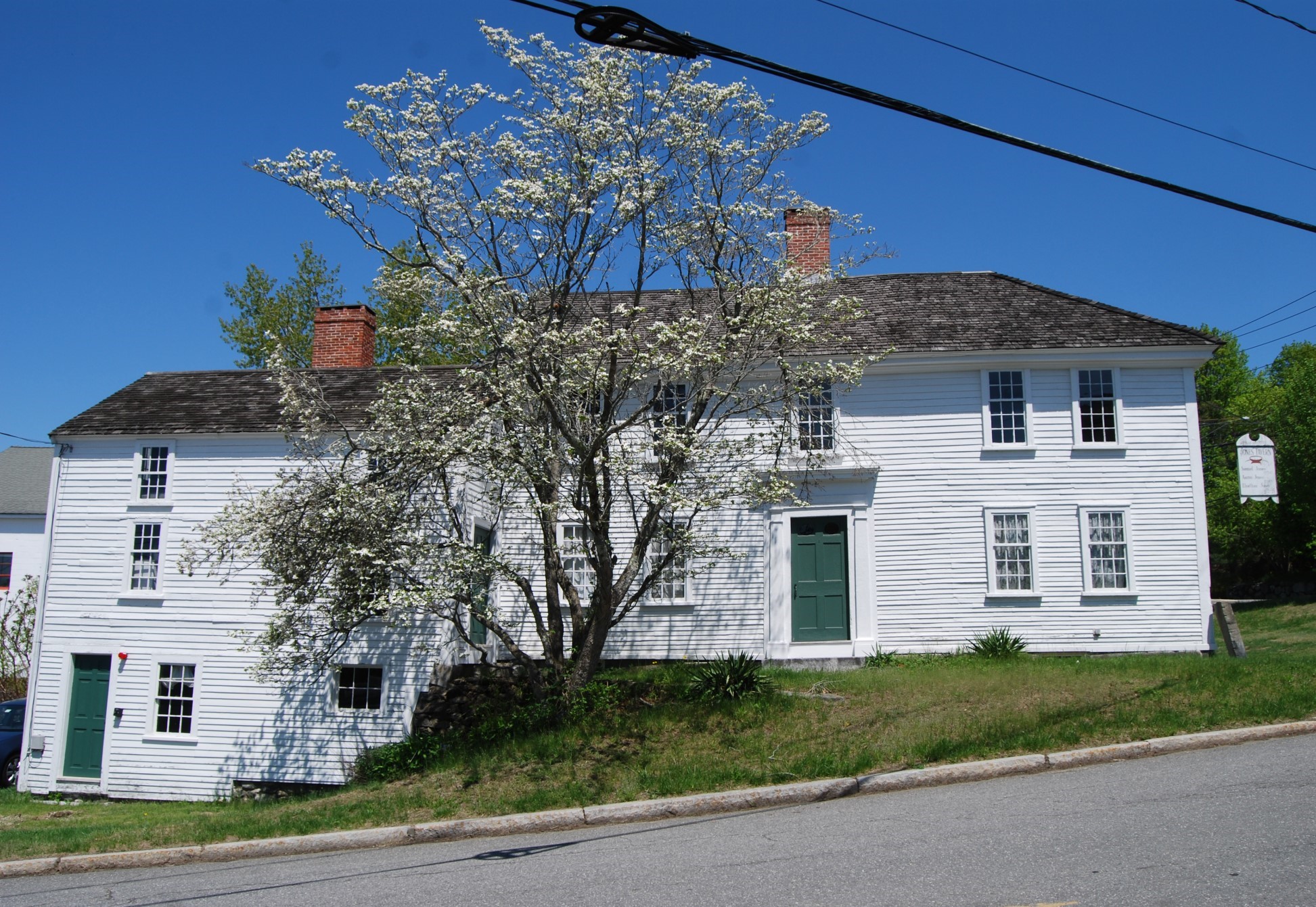 A Revolutionary-era white house with a center chimney. The main house is set on a hill and an ell on the left side has a small door that opens at a lower level.