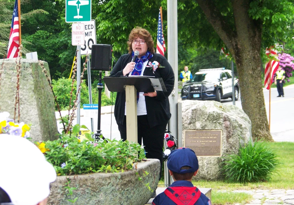 A woman wearing a black suit and a boutonniere with tri-color ribbon speaks at a podium at the square in West Acton.