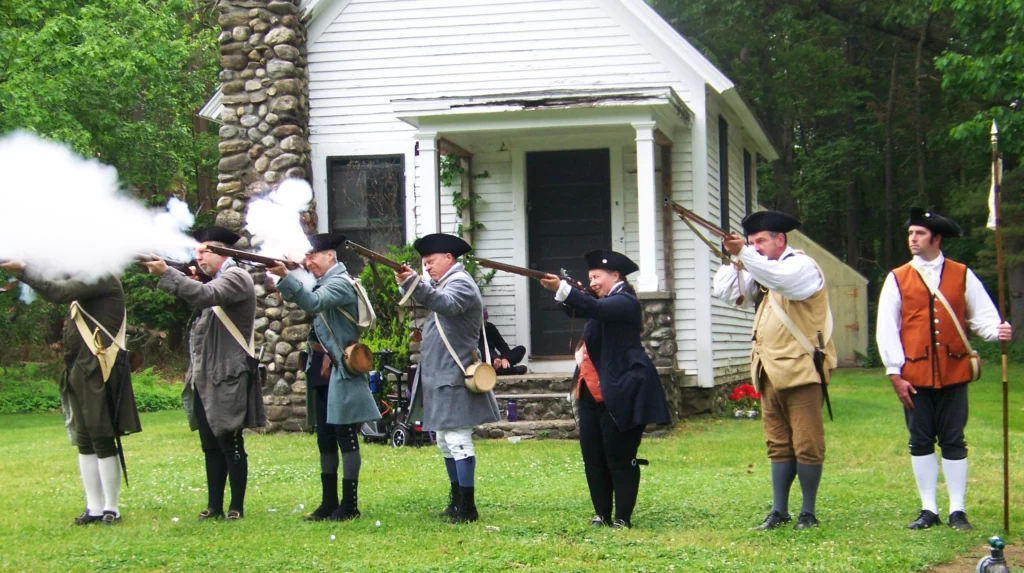 A line of Minutemen (and women) fire a volley of muskets in front of a small white wood and stone building.