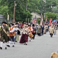 A group of men and women in Colonial garb are walking down a street in West Acton. The AB Marching Band is coming up behind them. There are folks walking alongside the parade and more watching from the sidewalk