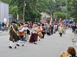 A group of men and women in Colonial garb are walking down a street in West Acton. The AB Marching Band is coming up behind them. There are folks walking alongside the parade and more watching from the sidewalk