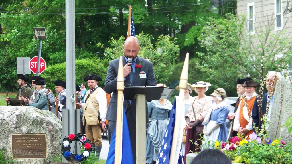 A bald man wearing a suit speaks at the podium in the West Acton square. The Minutement are in the background and some residents are watching. There are several flags in the picture.