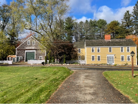 A yellow center-chimney house with a large, old-fashioned barn on the left.