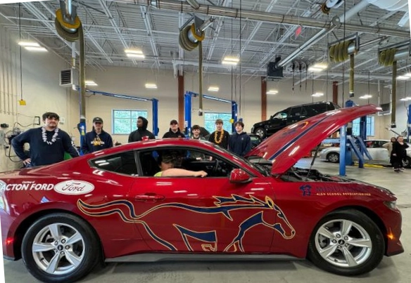 A side view of a brand new, bright red, Ford Mustang with a special paint job that includes a picture of a running horse and both Acton Ford and Minuteman Tech logos.