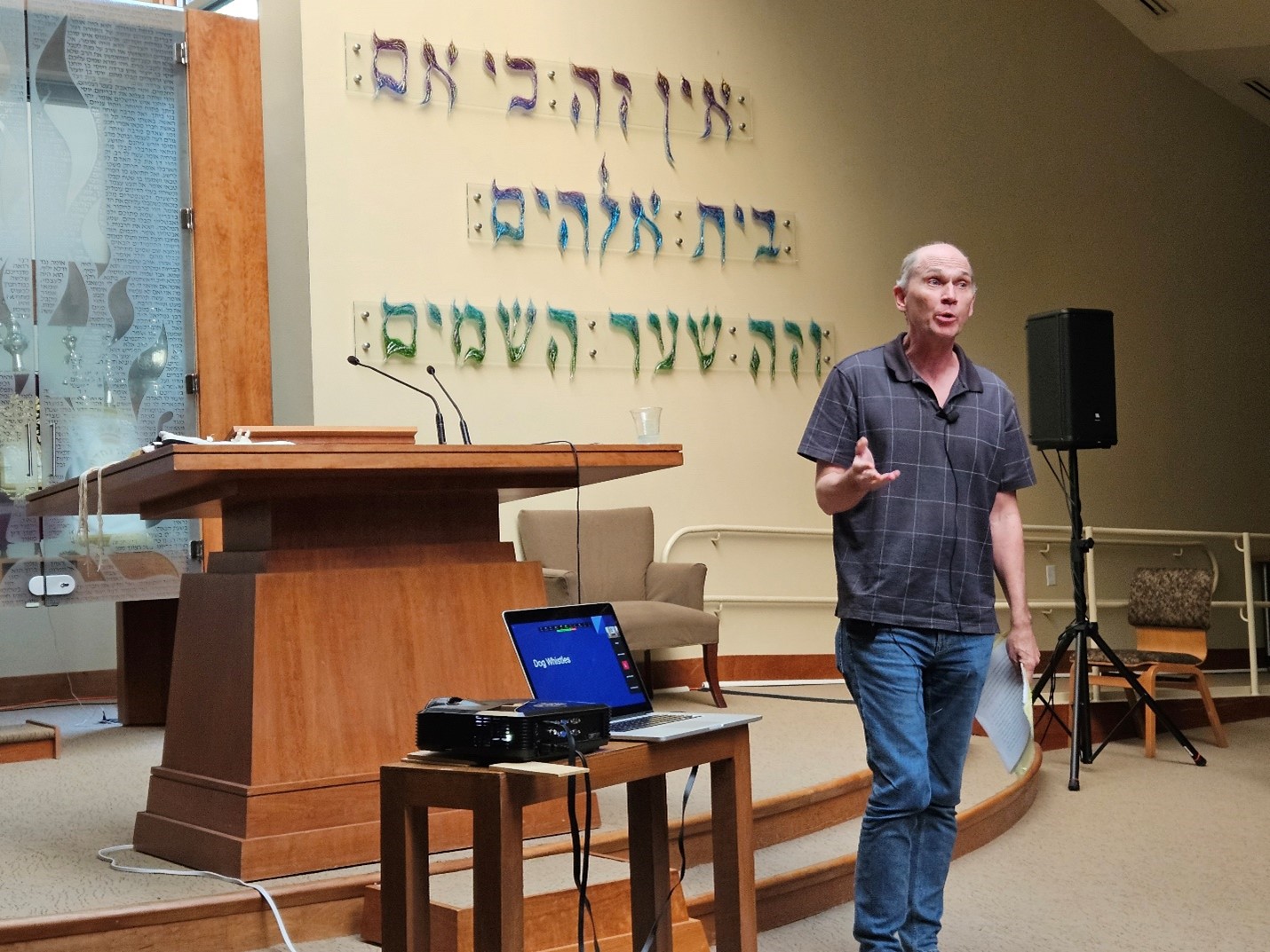 A tall grey-haired man wearing a polo and jeans stands at the front of the synagogue. A laptop sits on a table next to him, and Hebrew text is on the wall behind him.