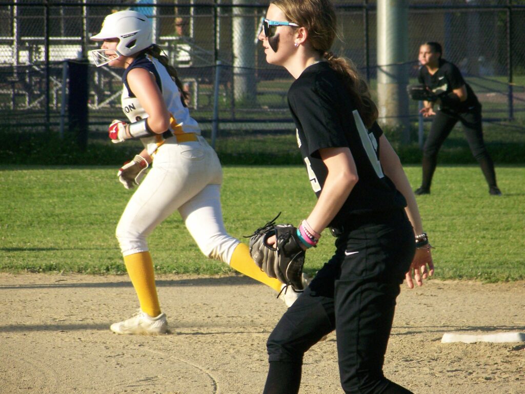 Two young women from opposing teams look intently towards home plate. The Revs player is on the base, waiting for the next hit, and the Springfield player is ready to protect the base.